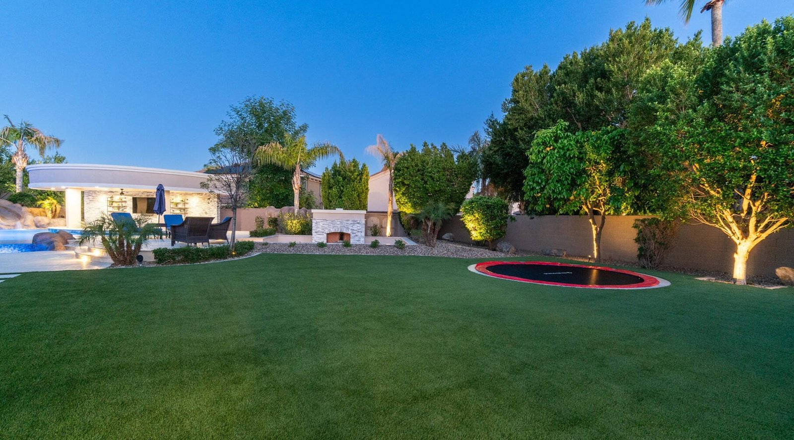 A spacious backyard with a perfect-looking artificial lawn, in-ground trampoline, trees, and landscaped plants. The area features a stone fireplace, lounge chairs, and umbrellas near an inviting swimming pool. The sky is clear, indicating it's either morning or evening.