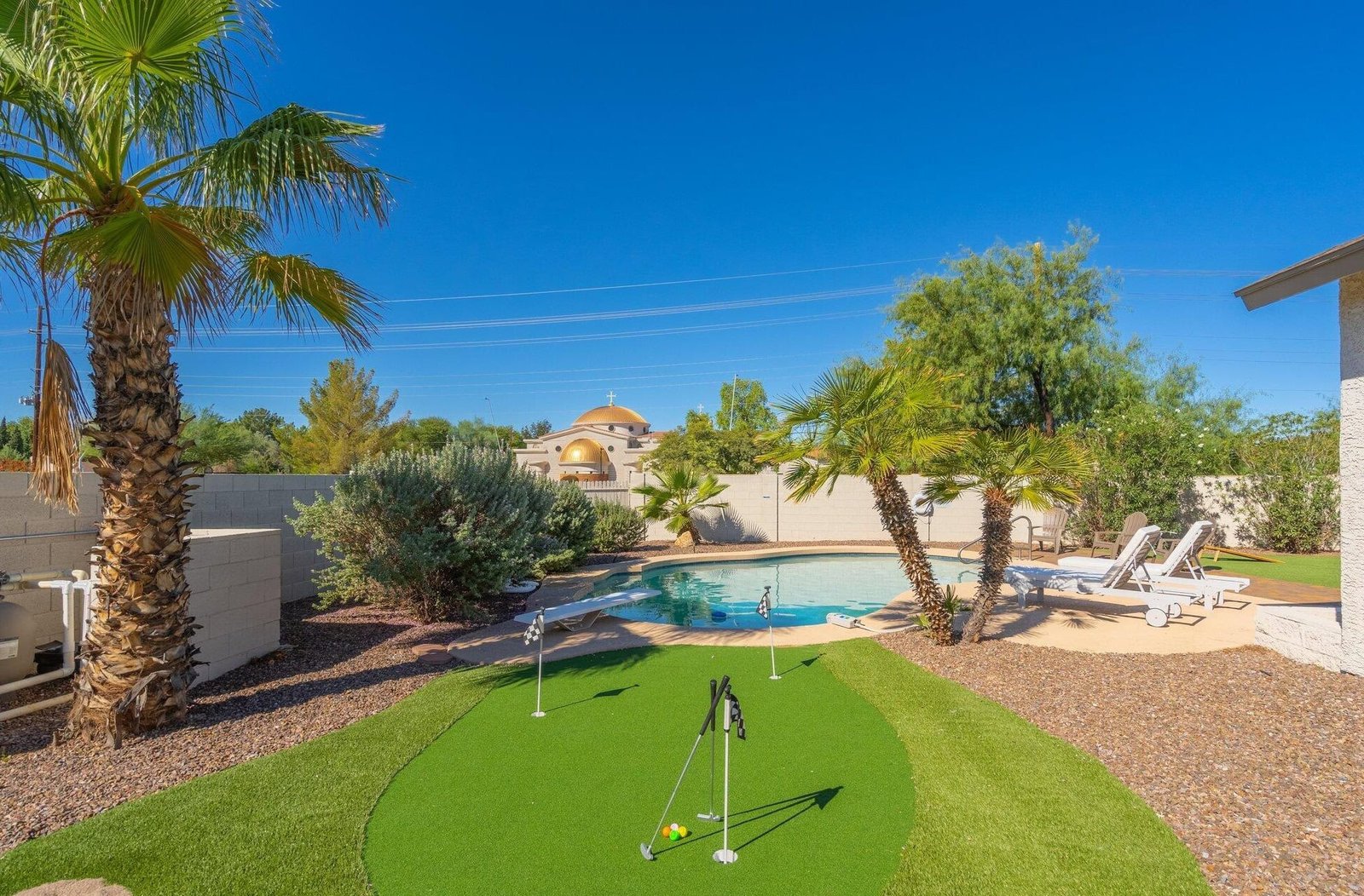 A backyard in Peoria, Arizona featuring a mini putting green with pristine artificial grass installation in the foreground. A small, curved pool surrounded by concrete sits in the middle, with multiple reclining chairs beneath an umbrella on the far side. Palm trees and desert foliage complete the scenic yard. Contact us for a free quote!