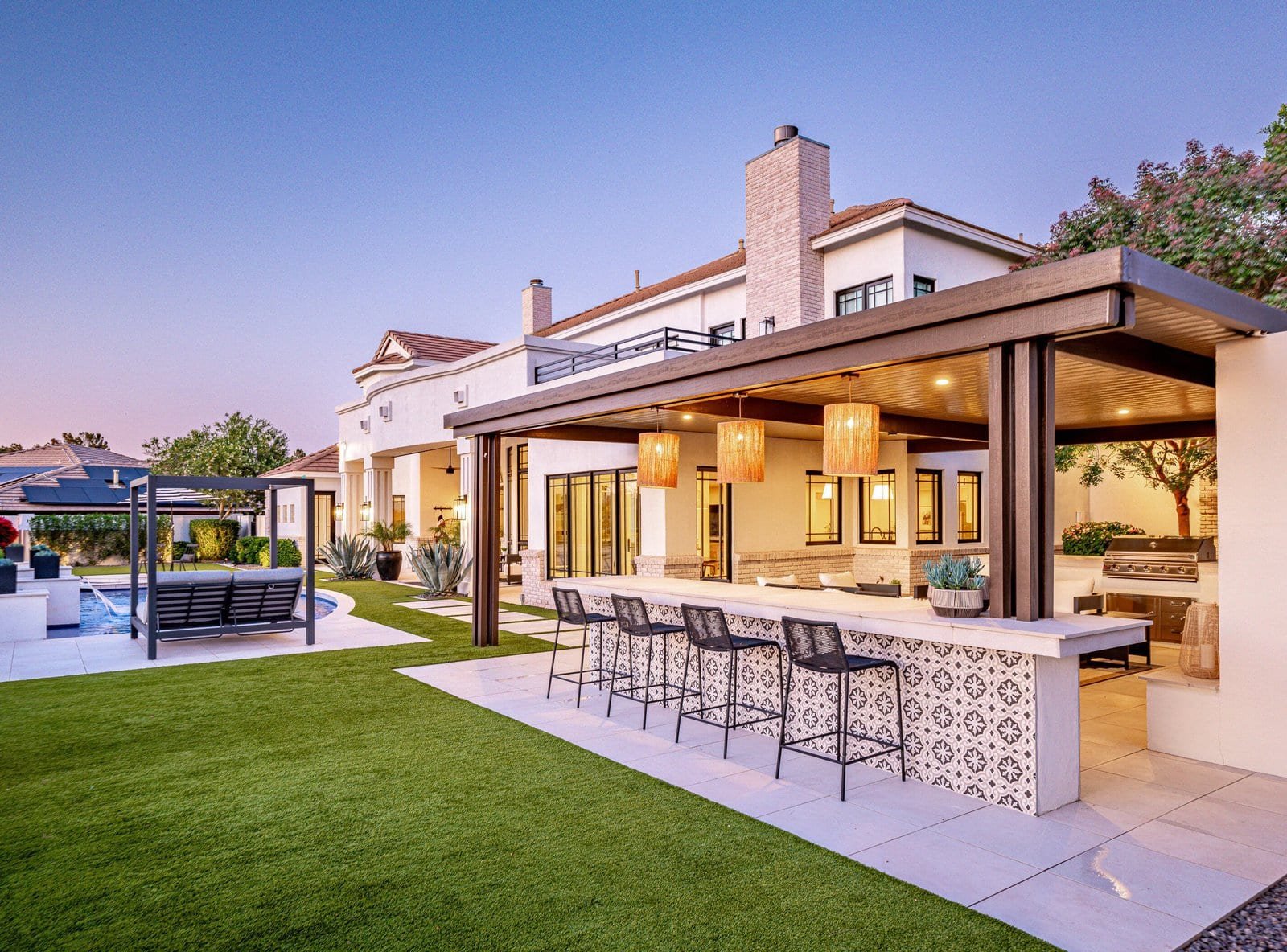 A luxurious backyard in Peoria featuring a large house, an outdoor kitchen with a tiled bar and stools, a covered patio with wooden accents and pendant lights, a pool, a lounge area with a swing, manicured green lawns adorned with various plants and trees, and premium artificial turf for easy maintenance.