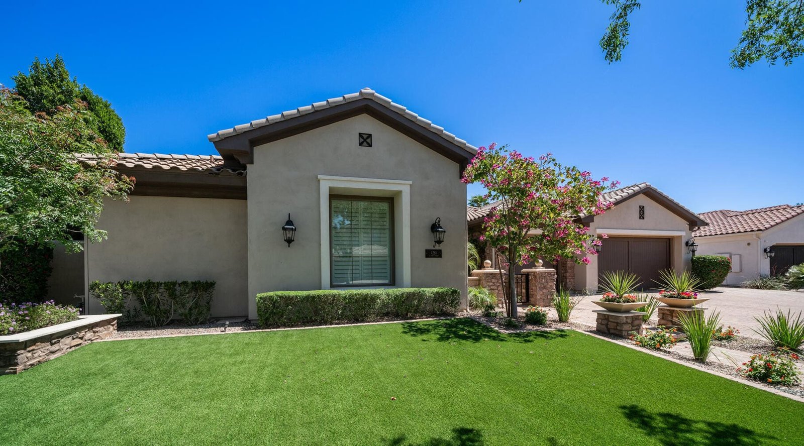 A single-story house in Peoria with beige stucco walls, a tiled roof, and a well-manicured lawn featuring premium artificial turf. The entrance has a window with white shutters and is flanked by sconces. A tree and various plants adorn the front yard, while the driveway leads to a two-car garage.