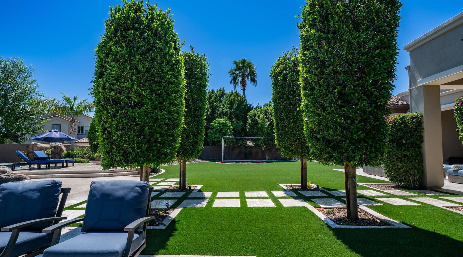 A well-manicured backyard in Peoria, Arizona features tall, neatly trimmed trees lining a path of stone pavers set in lush green grass. Lounge chairs to the left and a house with a patio to the right complete the scene. Considering artificial grass installation? Get a free quote today!