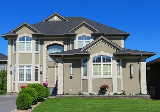 A two-story, modern beige house with dark gray roofing, large windows, and decorative pillars, located in Peoria Arizona. The front yard features a well-maintained lawn with artificial grass installation, small bushes, and flowerbeds. The sky is clear and blue, suggesting a bright, sunny day.