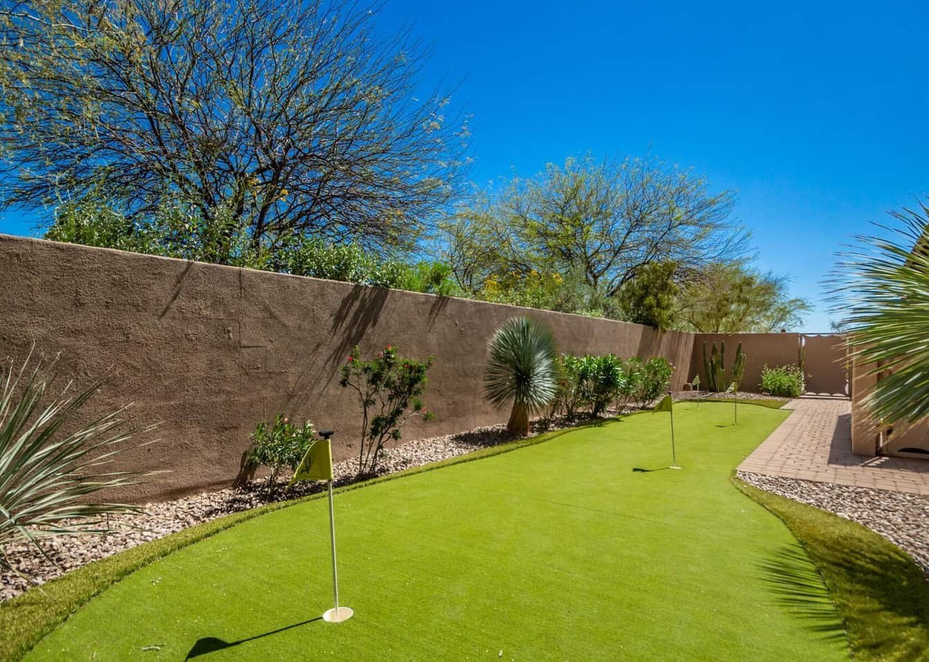 A backyard scene in Peoria featuring a well-maintained putting green with several golf holes and flags on artificial turf. Surrounding the green are desert plants, including trees and shrubs, bordered by a stucco wall. The sky is clear and bright blue, indicating a sunny day.